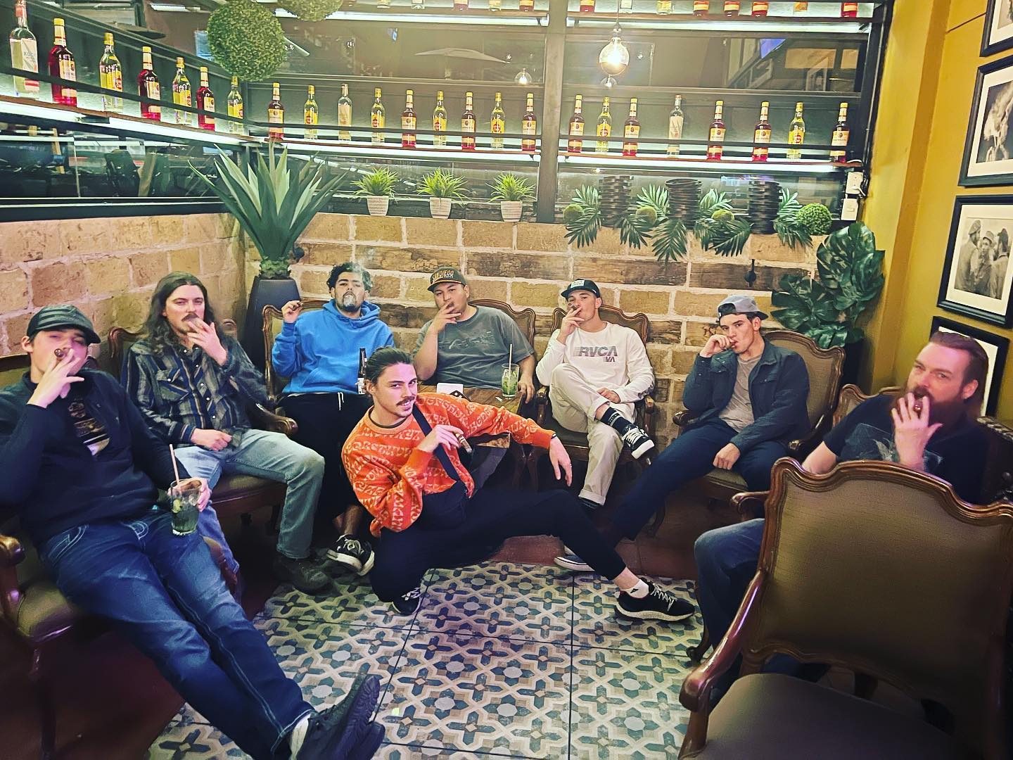 A group of friends sitting together indoors, several holding cigars
